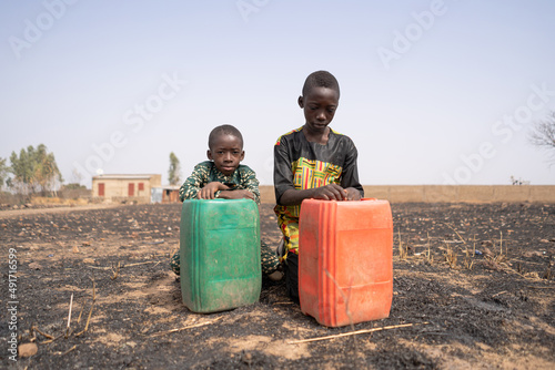 Two sad African boys kneeling in a burnt field behind their large water canisters, staring desperately at the ground in front of them; Water shortage icon