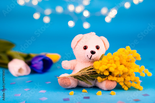 Cartolina di Buon Compleanno con un orsacchiotto bianco che tiene in braccio un mazzetto di mimose e nello sfondo tre tulipani colorati su sfondo azzurro con luci sfuocate. Auguri. Anniversario. 