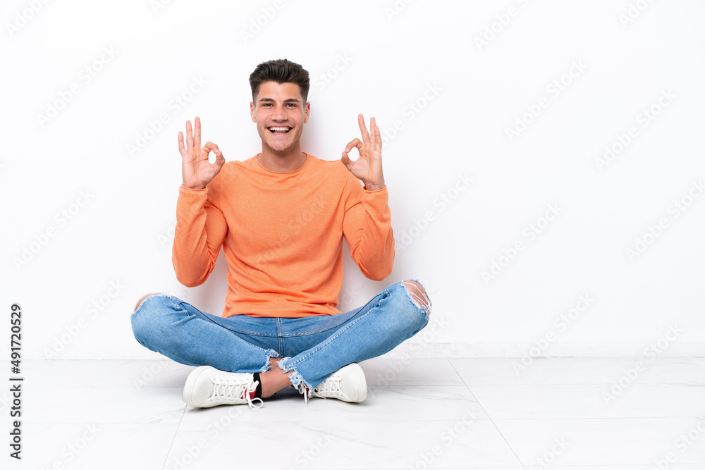 Young man sitting on the floor isolated on white background showing an ok sign with fingers Young man sitting on the floor isolated on white background showing an ok sign with fingers