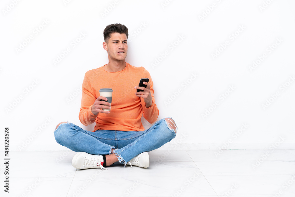 Fototapeta premium Young man sitting on the floor isolated on white background holding coffee to take away and a mobile while thinking something