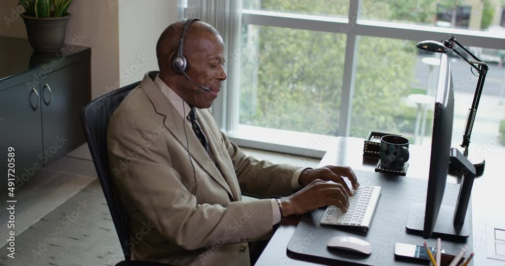 Side profile view of a happy corporate black business man typing on a desktop computer while talking on a headset near a large office window. 35mm Medium Closeup Tripod Shot 4K.
