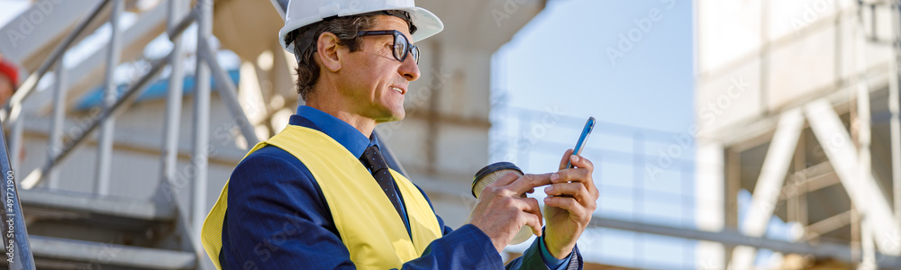 Matured man in work vest holding cup of coffee and texting message on smartphone Matured man in work vest holding cup of coffee and texting message on smartphone