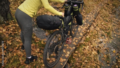 The woman travel on mixed terrain cycle touring with bikepacking. The traveler journey with bicycle bags. Sportswear in green black colors. The trip in magical autumn forest. Wooden bridge, river.