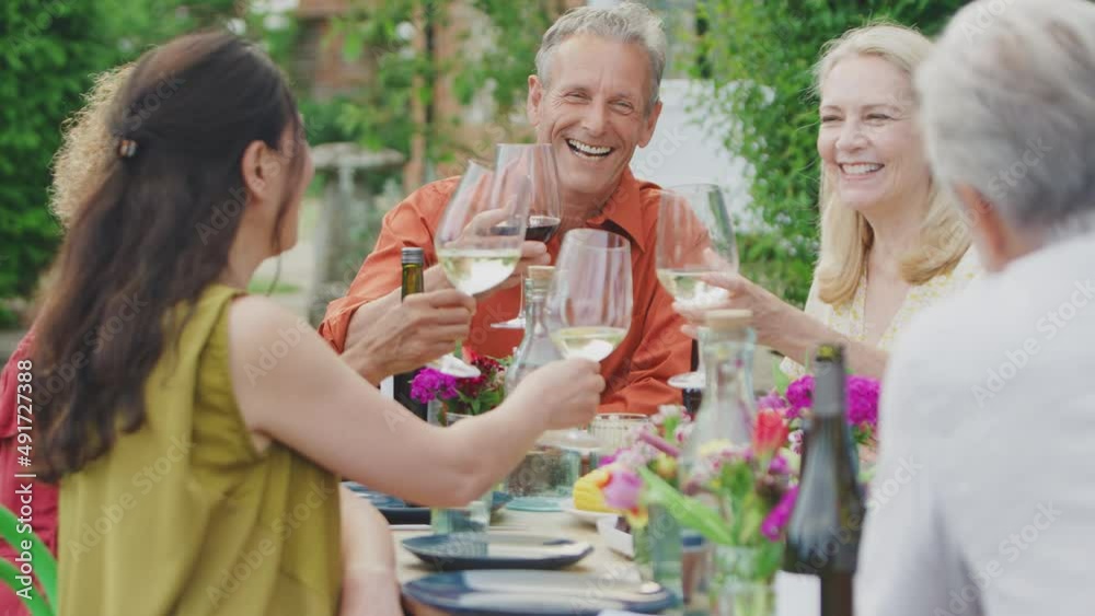 Group of mature friends sitting around table talking and making a toast with wine at summer drinks party in garden at home - shot in slow motion Group of mature friends sitting around table talking and making a toast with wine at summer drinks party in garden at home - shot in slow motion