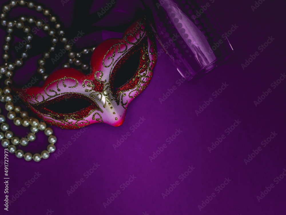 A masquerade mask, white pearl beads and an empty champagne glass on a purple-lilac background. A masquerade mask, white pearl beads and an empty champagne glass on a purple-lilac background.
