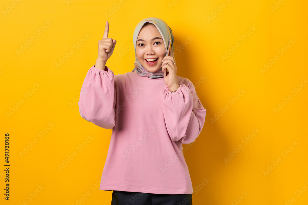 Smiling young Asian woman in pink shirt talking on mobile phone and pointing fingers up, having a good idea on yellow background Smiling young Asian woman in pink shirt talking on mobile phone and pointing fingers up, having a good idea on yellow background