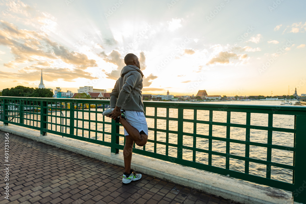 Fototapeta premium Confidence African man in sportswear stretching his body while jogging exercise on the bridge in the city at summer sunset. Strong male athlete enjoy outdoor activity sport training running workout
