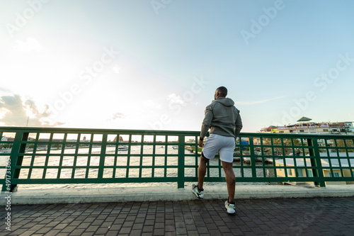 Fototapeta Naklejka Na Ścianę i Meble -  Confidence African man in sportswear and smart watch jogging exercise on the bridge in the city at summer sunset. Strong male athlete enjoy outdoor activity lifestyle sport training running workout. 