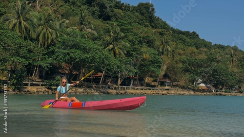 Young blonde woman in blue swimsuit rows pink plastic canoe along azure sea bay past island with palms under blue sky at resort. Traveling to tropical countries. Girl is sailing on kayak in ocean. © ivandanru