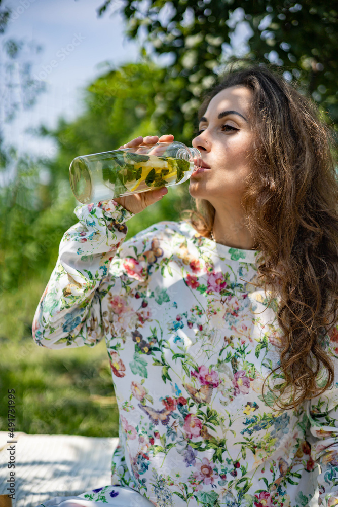 Fototapeta premium Portrait of beautiful woman posing lying on plaid at summer garden park. A book, fruit, a bag and a watermelon are next to each other.