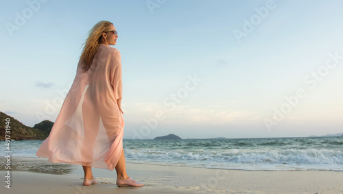 Sexy hot woman walks on golden sunset on tropical beach  wet sand  against background of sea. Girl in green swimsuit and pink tunic silk shirt cape  drinks her orange cocktail Pina Colada.