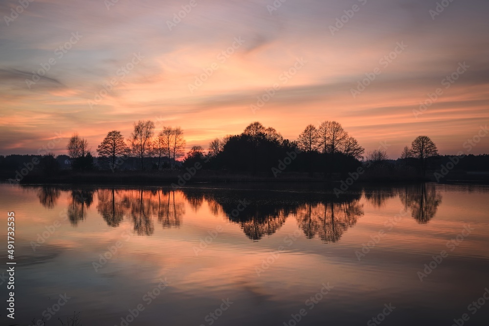Beautiful morning landscape by the water. Wonderful sunrise over the lake with the reflection of trees.