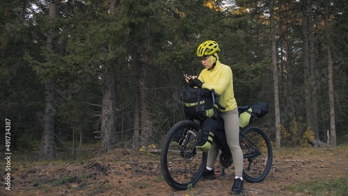The woman travel on mixed terrain cycle touring with bike bikepacking outdoor. The traveler journey with bicycle bags. Sportswear in green black colors. Magic forest park. Make a selfie smartphone. © ivandanru