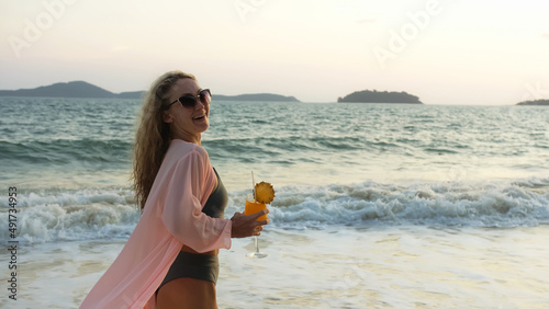 Sexy hot woman walks on golden sunset on tropical beach, wet sand, against background of sea. Girl in green swimsuit and pink tunic silk shirt cape, drinks her orange cocktail Pina Colada.