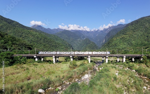A local train traveling on a bridge over the grassy, stony riverbed in Sanjhan valley with the mountaintops veiled by the clouds under blue sunny sky in Taroko National Park, Xincheng, Hualien, Taiwan