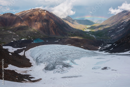 Bleak landscape with vivid rainbow above mountain lake. Gloomy scenery with bright rainbow above glacier and glacial lake in mountain valley. Top view to colorful rainbow and low clouds in mountains.