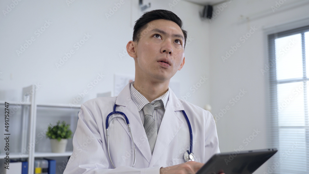 close up man in doctor’s uniform is pondering with hand on the chin while searching for information on his tablet computer in a bright workplace.