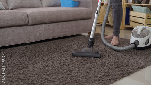 close up view vacuum cleaner head moving back and forth on the gray rug by the sofa with a woman standing barefoot in the background.
