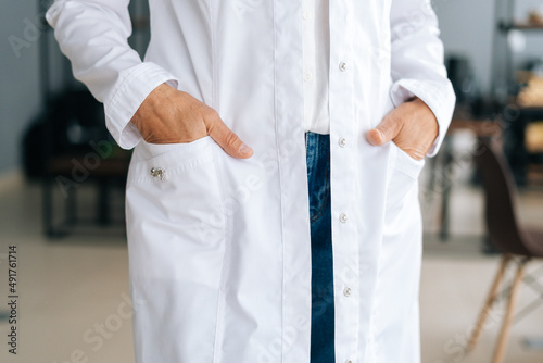 Close-up cropped shot of unrecognizable female doctor in white uniform holding hands in pockets standing in hospital office. Practitioner woman posing at workplace. Concept of medical work.