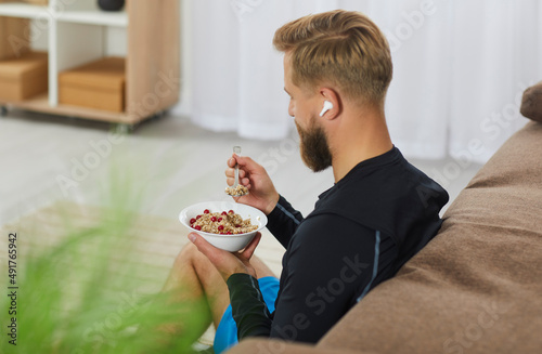 Healthy lifestyle. Athletic millennial man starts his day with healthy oatmeal with berries for breakfast. Unrecognizable man with wireless headphones in his ears sitting on sofa with bowl of oatmeal.