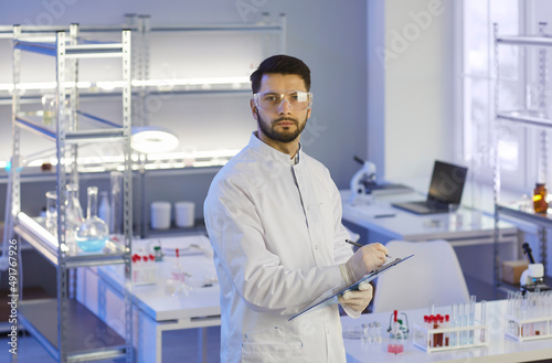 Portrait of serious researcher conducting scientific research in modern medical laboratory. Young attentive man in white coat stands in interior of laboratory with clipboard and looks into camera.