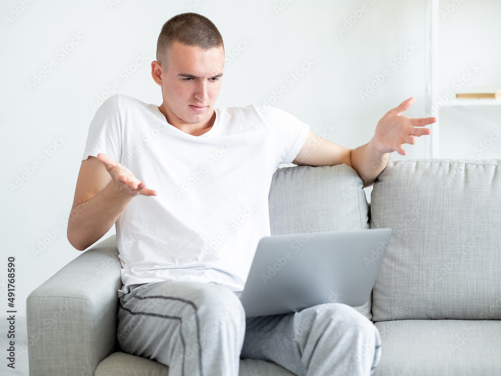Uncertain situation. Hesitating man. Online meeting. Confused casual guy sitting sofa looking laptop with shrugging hands in light room interior.