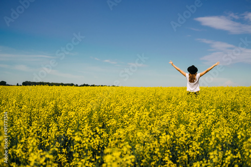 the woman in the hat enjoying nature in the middle of field full of yellow flowers. Yellow field of flowering rapeseed with cloudy blue sky - brassica napus - plant for green energy, medicine.