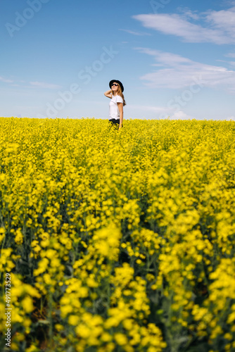 the woman in the hat enjoying nature in the middle of field full of yellow flowers. Yellow field of flowering rapeseed with cloudy blue sky - brassica napus - plant for green energy, medicine.