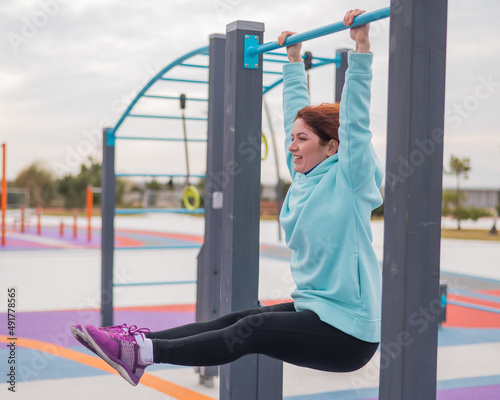 Caucasian woman in a mint sweatshirt hangs on a horizontal bar and does an exercise of the abdominal muscles on the sports ground outdoors. 