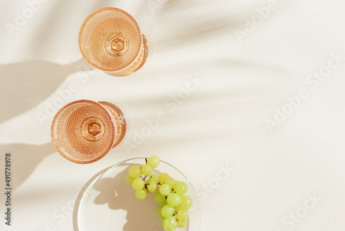 Two glasses of white wine from pale pink colored glass with palm leaf shadow from sunlight and plate with grapes fruit, summer drink minimal concept, modern glass on peach color background