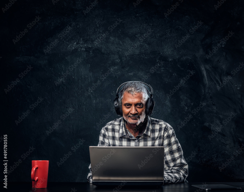 Modern old man with laptop sitting at table against dark background Modern old man with laptop sitting at table against dark background