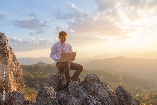 Businessman working outdoors with laptop computer on top rock mountain at sunset background. Work and vacation.