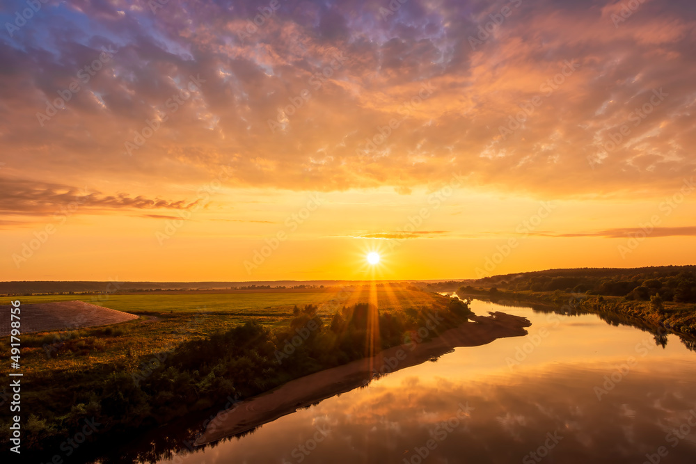 Scenic view at beautiful sunset or sunrise on a shiny river with green bushes on sides, golden sun rays, calm water ,deep blue cloudy sky and forest on a background, spring landscape