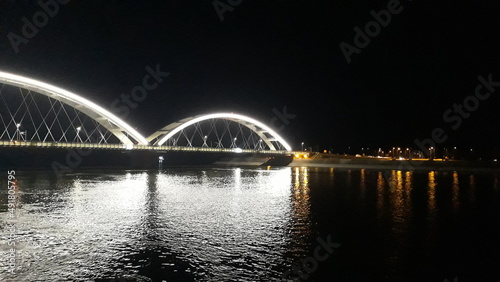 Die Brücke über den Fluss wirft Weiß in die Nacht. Die Lichter der nächtlichen Stadt spiegeln sich im Fluss. Silhouetten von Siedlungen am See. Verschwommene Fotos in der Dunkelheit der Nacht.