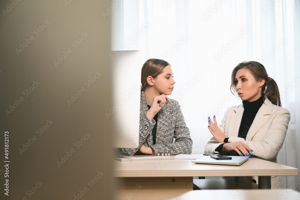 Attractive young adult women work together in the office at a meeting. A new employee consults with a mentor and presents a new project. Open space concept for teamwork Attractive young adult women work together in the office at a meeting. A new employee consults with a mentor and presents a new project. Open space concept for teamwork