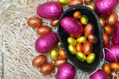 Pile of colorful foil wrapped chocolate easter eggs in pink, red, silver and gold with two halves of a large brown dark chocolate egg in the middle and mini eggs inside, on pale shreaded cream nest. photo