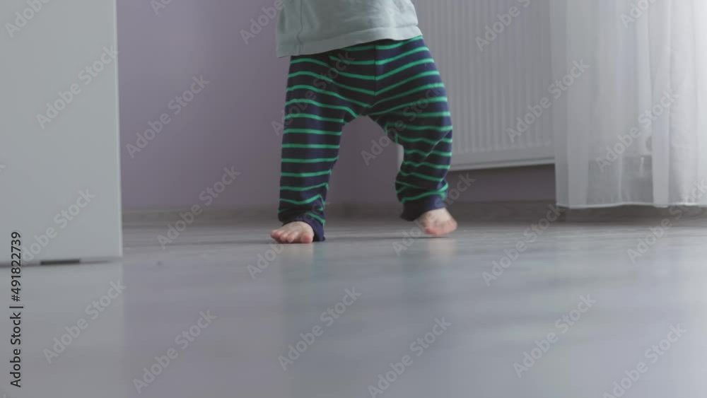 unrecognizable baby in striped pants making first steps barefoot on grey floor in living room indoors, toddler learning to walk indoors, child kid development physical skills. walking home bare feet unrecognizable baby in striped pants making first steps barefoot on grey floor in living room indoors, toddler learning to walk indoors, child kid development physical skills. walking home bare feet