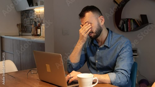 Man with beard working on computer. Sitting in home typing on laptop tired takes off glasses and rubbing nose and eyes feeling fatigue and headache. Stress and frustrated concept