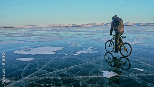 Man is riding bicycle on the ice. Ice of the frozen Lake Baikal. Rider is dressed in black down jacket, cycling backpack, helmet. Tires on covered with special spikes. Traveler boy is ride cycle.