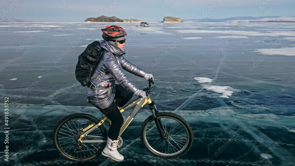 Fototapeta premium Woman is riding bicycle on the ice. Girl is dressed in a silvery down jacket, cycling backpack and helmet. Ice of the frozen Lake Baikal. Tires on bike are covered with spikes. Traveler is ride cycle.