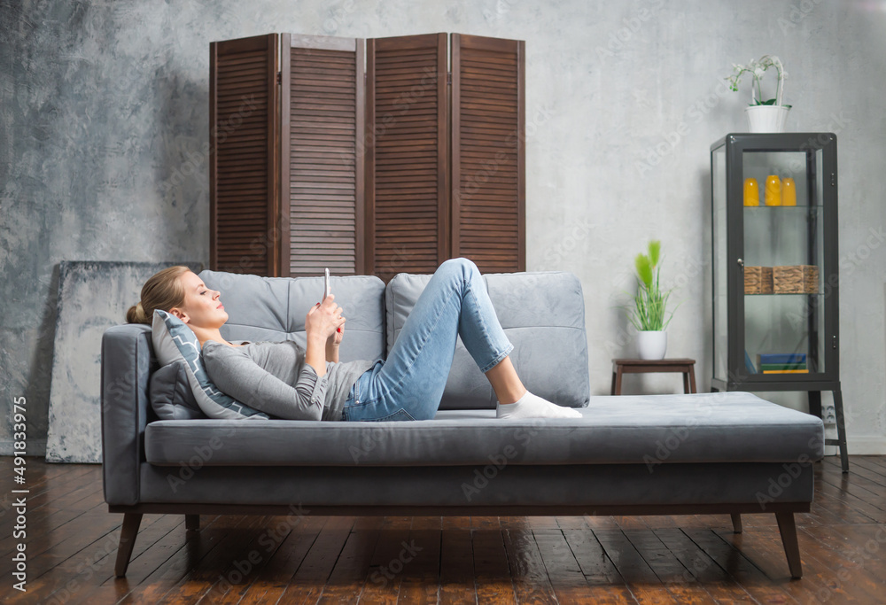 Young woman works using a smartphone lying on the couch. Student, entrepreneur or freelancer girl working or studying remotely via internet. Young woman works using a smartphone lying on the couch. Student, entrepreneur or freelancer girl working or studying remotely via internet.