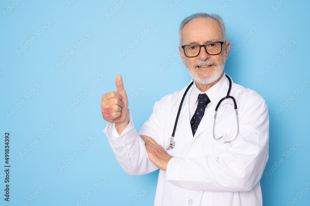 Senior grey-haired doctor man standing over isolated blue background happy face smiling with thumb up looking at the camera. Positive person. Senior grey-haired doctor man standing over isolated blue background happy face smiling with thumb up looking at the camera. Positive person.