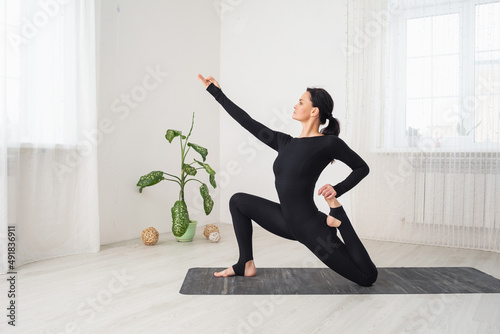 Woman in black sportswear practicing yoga doing anjaneyasana exercise with virasana, low lunge pose, sitting on a mat in a room by the window