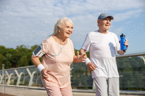 Senior gentleman with cap and bottle and woman with gadgets run along footbridge