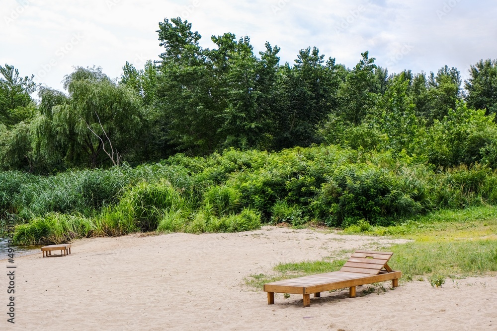Wooden deck chair on the beach against the backdrop of greenery on the banks of the river. Summer recreation calm peaceful area. Tourism and traveling, vacations and outdoor activity. Relaxation.