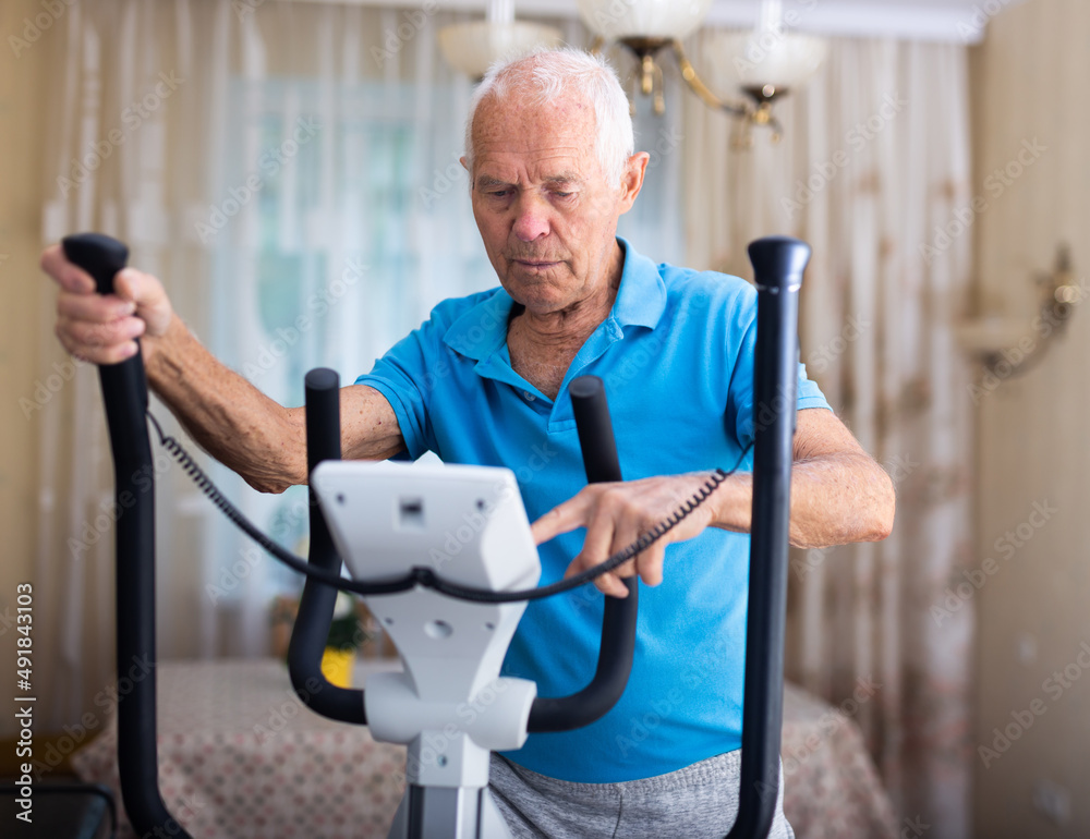 Positive senior man works out on an elliptical machine at home Positive senior man works out on an elliptical machine at home