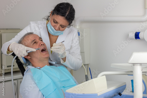 Middle-aged man is sitting on dentist chair with mouth opened while dentist is working on his ill tooth.