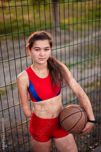 Young woman athlete with an amputated arm and burns on her body. She hold in the hands basketball ball after training outdoor at sunset. © artem_goncharov