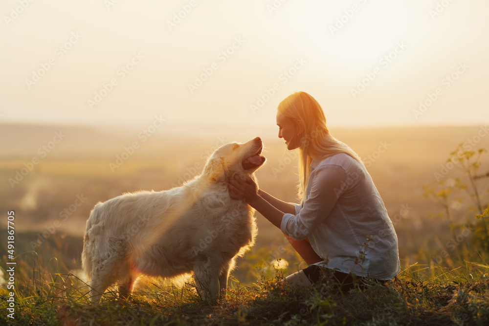 Happy young woman is sitting on the hill at sunset lovingly hugging her large breed dog.