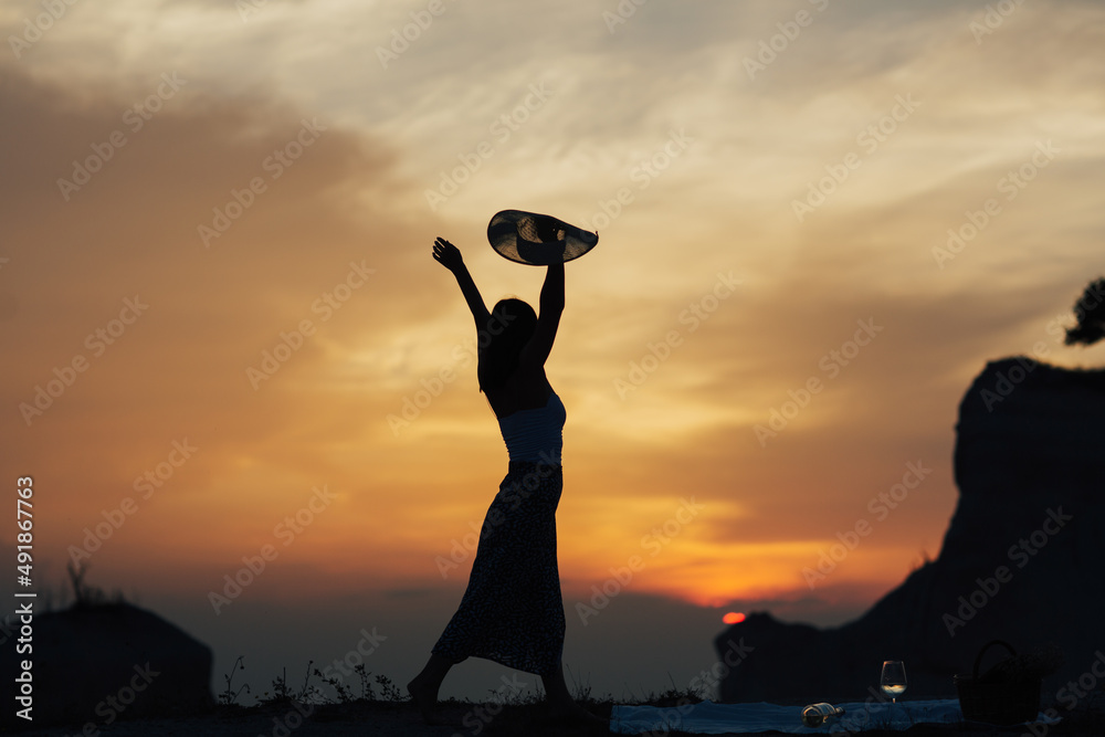 Silhouette of young female with hands raised standing high up on a mountain at sunset.